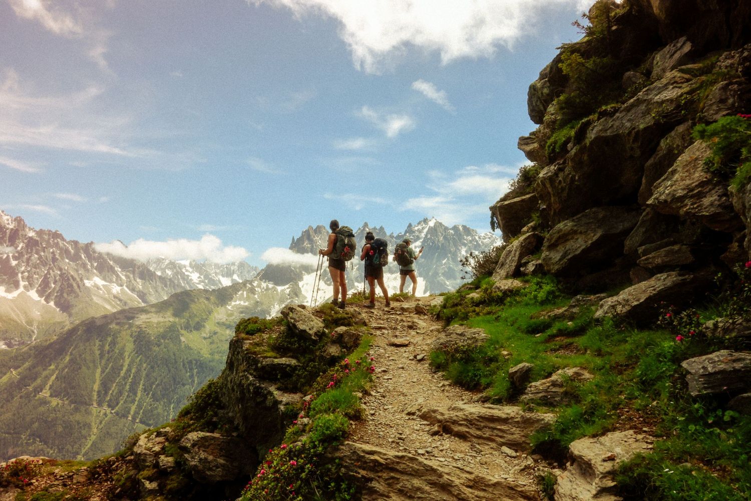 Three hikers on a rocky trail high above an alpine valley, with snowy peaks behind them