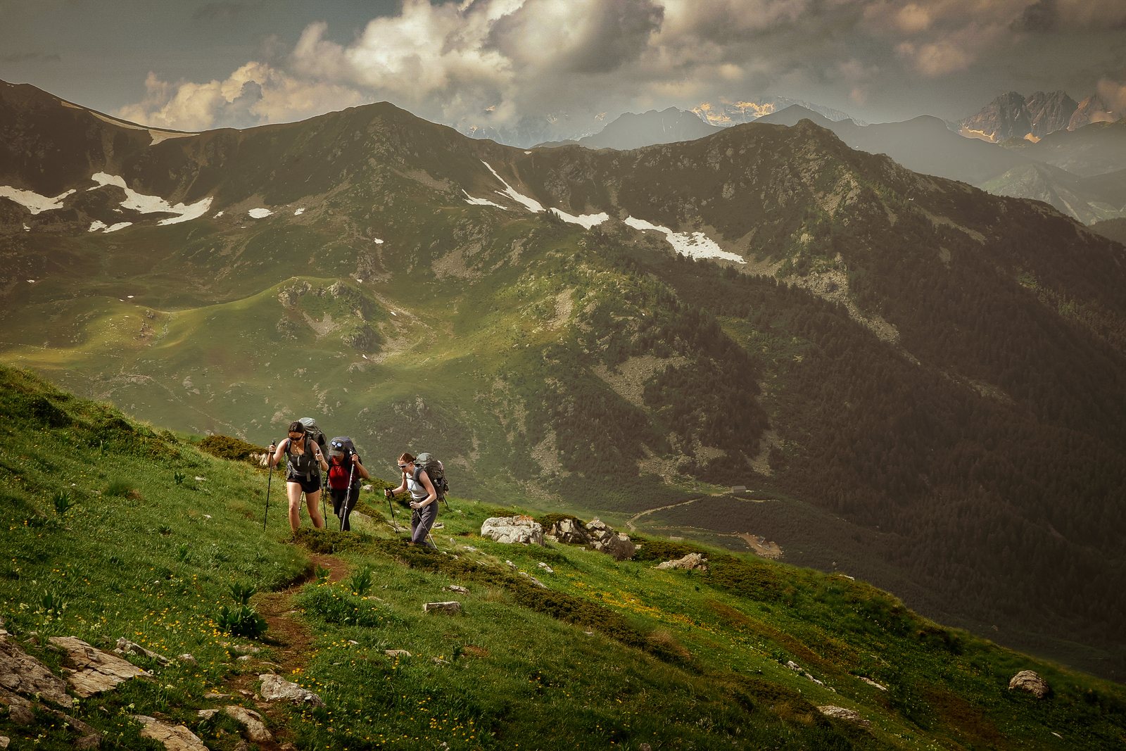A line of hikers on a green ridge below snowy mountains
