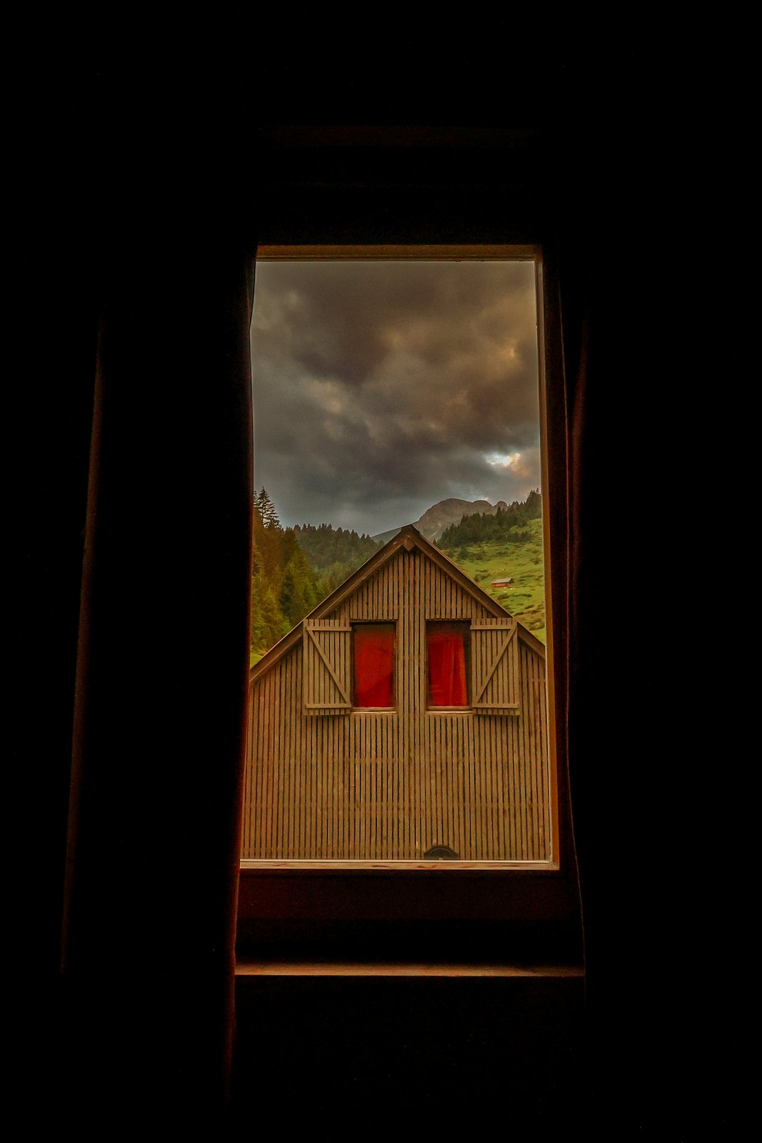 A dark hut window framing a wooden cabin under storm cloud