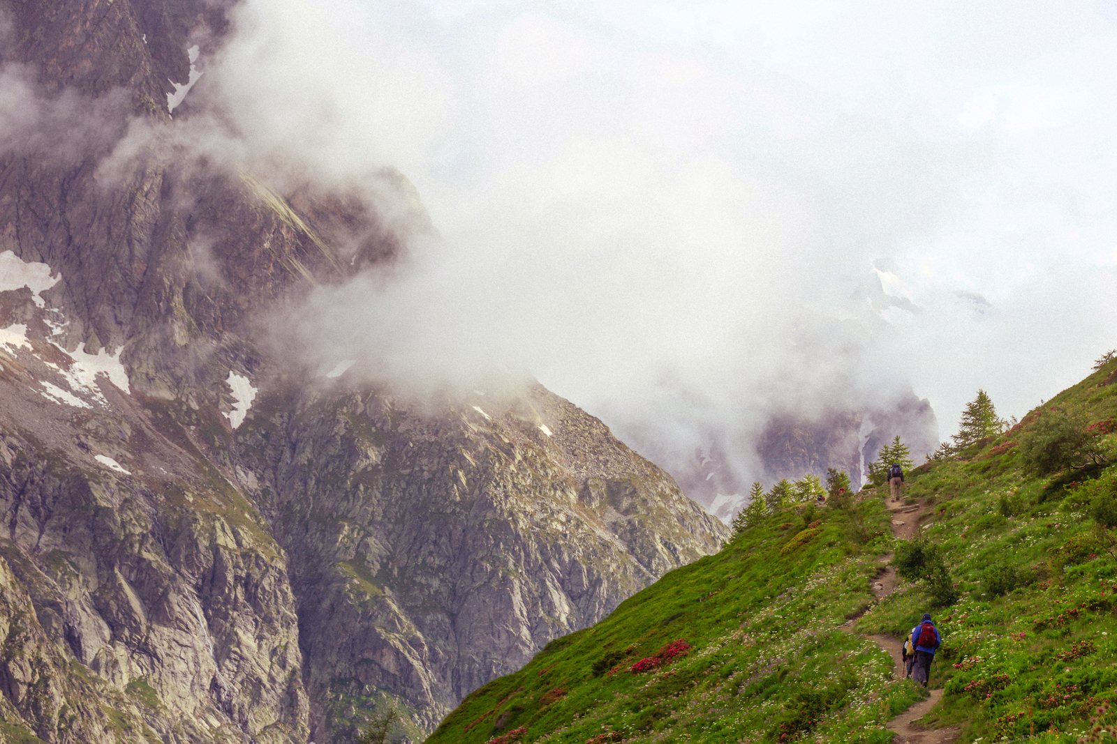 A lone hiker on a green slope below cloud-wrapped cliffs